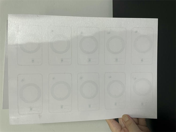 Ръка държи HF RFID преламинационен инлей лист показващ пръстеновидни антени вътре в правоъгълни контури на карти.